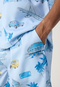 Chemise à manches courtes en tissu bleu clair avec des illustrations de voitures, de palmiers et de monuments dans des nuances de bleu et de noir.