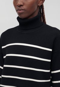 Persona che indossa un maglione nero a collo alto con strisce orizzontali bianche e capelli intrecciati su uno sfondo chiaro uniforme.