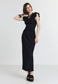 Robe noire avec un tissu texturé, des manches volantées, un corsage ajusté et une longue jupe. Associée à des sandales à plateforme et un collier superposé.
