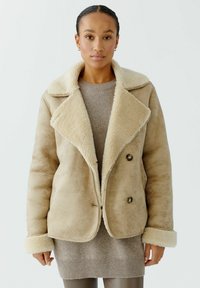 Beige schapenvachtjas met grote, zachte kraag, dubbelknopen en pluche manchetten, gedragen over een lichte trui en strakke leggings.