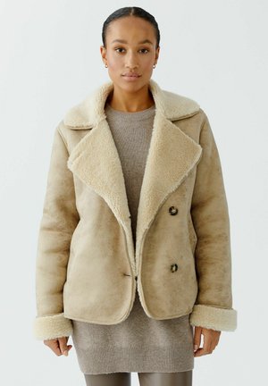 Manteau en shearling beige avec un large col doux, des boutons croisés et des poignets en peluche, porté sur un pull léger et un legging ajusté.