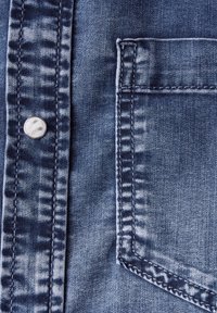 Tessuto denim blu con una leggera sfumatura, dotato di un bottone metallico e una tasca sul petto. Primo piano che mostra i dettagli delle cuciture.