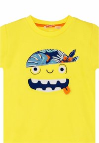 Camisa amarilla con un rostro de dibujos animados que lleva un pañuelo azul con estampado, ojos grandes, dientes cosidos y un detalle de un helado de naranja.