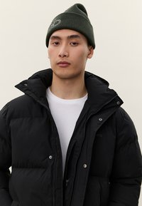 Jauns vīrietis ar olīvu zaļu cepuri, baltu kreklu un melnu pūkainu jaku uz vienveidīga gaiša fona.