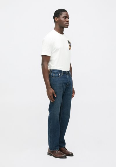 Valge puuvillane t-särk graafilise taskudisainiga, kombineeritud tumeliste siniste teksapükstega. Komplektis tumepruunid kingad. Vabaaja riietus, külgvaade.