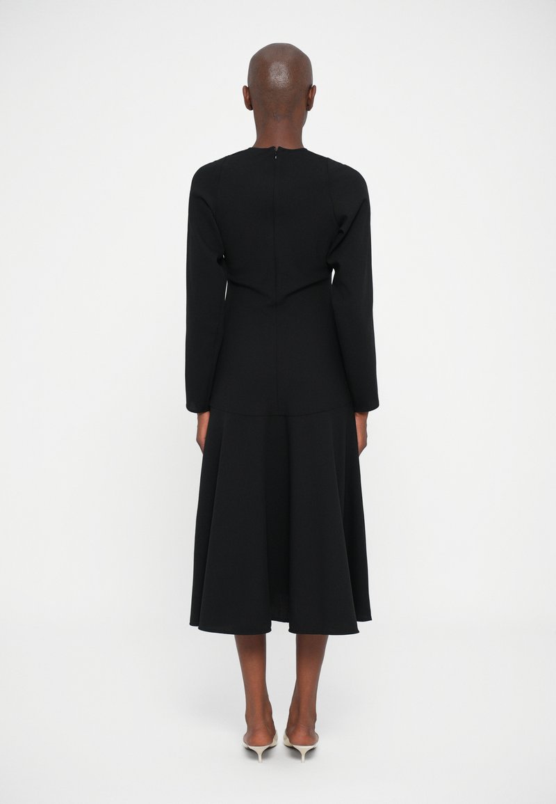 Personne avec la tête rasée portant une robe midi noire à manches longues et des escarpins blancs à bout pointu, se tenant face à un fond blanc.