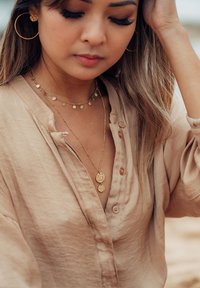 Beige Hemd mit Knöpfen und einer subtilen Textur, kombiniert mit geschichteten goldenen Halsketten mit runden Charme-Akzenten und großen goldenen Creolen.