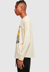 Beige Langarm-T-Shirt mit einem großen schwarzen und gelben grafischen Design auf der Vorderseite. Runder Ausschnitt und lockere Passform. Getragene schwarze Hose.