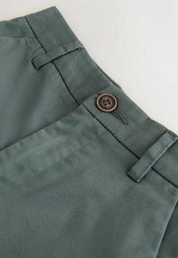 Cinturilla de pantalones verdes con trabillas para cinturón y un botón marrón etiquetado como "Authentic Dude Lyon", mostrando el bolsillo delantero y detalles de costura.