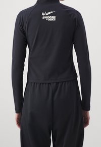 Svart atletisk jacka med långa ärmar, kort design. Har en hög krage och "Swoosh by Nike"-logotyp i vitt på baksidan. Slät textur.