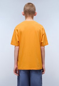 Kurzärmliges, Rundhals-T-Shirt aus leuchtend orangefarbenem Baumwollstoff. Die Rückansicht zeigt eine lockere Passform und keine Muster. Kombiniert mit dunkelblauen Cargo-Shorts.