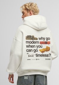 Sweat à capuche blanc avec un grand motif au dos présentant divers designs de meubles et le texte "pourquoi opter pour le moderne quand vous pouvez choisir l'intemporel ?"