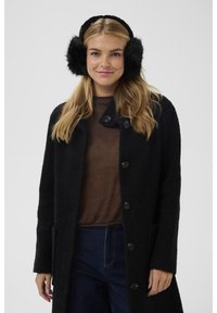 Jeune femme portant des cache-oreilles noirs en fourrure, un manteau texturé noir, un pull marron et un jean bleu foncé, souriante devant un fond gris uni.