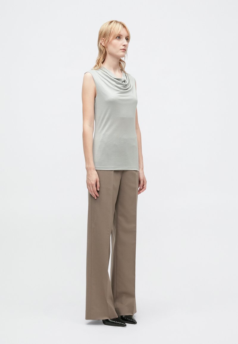 Top senza maniche grigio chiaro con scollo drappeggiato, abbinato a pantaloni beige a gamba larga con vita alta e scarpe nere a punta.