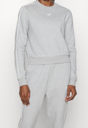 Personne portant un sweat-shirt Reebok gris clair et un pantalon de survêtement assorti avec un liseré blanc le long des manches et des jambes.