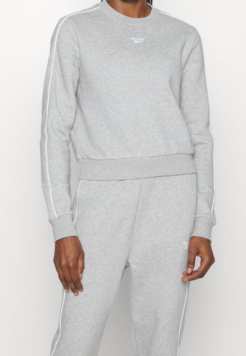 Personne portant un sweat-shirt Reebok gris clair et un pantalon de survêtement assorti avec un liseré blanc le long des manches et des jambes.