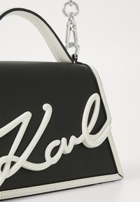Borsa a tracolla in pelle nera con dettagli bianchi e una vistosa firma bianca. Presenta una forma strutturata e una maniglia a catena argentata.