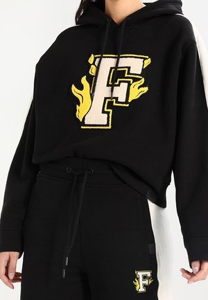 Person wearing black cropped hoodie and pants set with large white and yellow "F" and flame logo on chest and pants.