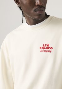 Hombre con sudadera crema con texto rojo "Levi Strauss & Company" bordado en el pecho izquierdo, mirando ligeramente hacia la derecha.