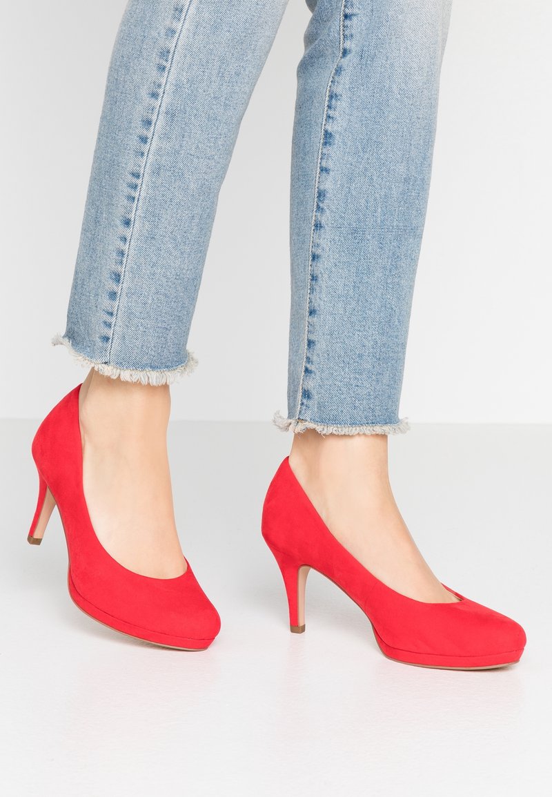 Escarpins à talons hauts en daim rouge avec un bout rond et un talon fin. Associés à un jean en denim bleu clair avec des ourlets effilochés.