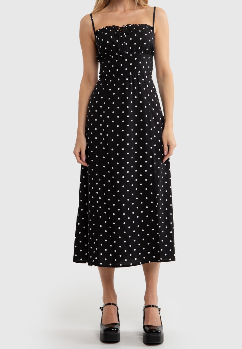 Kvinna iklädd en svart midiklänning med vita prickar och tunna axelband, kombinerat med svarta plattform Mary Jane-skor på en enfärgad bakgrund.