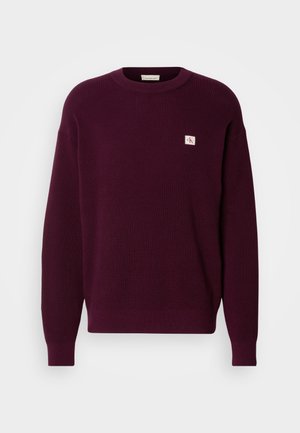 Burgundska pletena pulover s rebrastim vzorcem, krojem s krožnim izrezom in majhnim logotipom na prsih. Dolgi rokavi z rebrastimi manžetami.