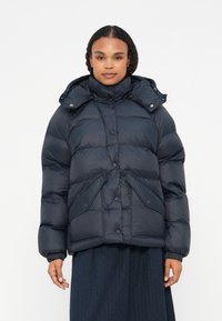 Mornarska puffer jakna z visokim ovratnikom, gumbnimi zapiranji in dvema sprednjima žepoma. Ima elastične manšete in odstranljivo kapuco. Mehka, quilted tekstura.
