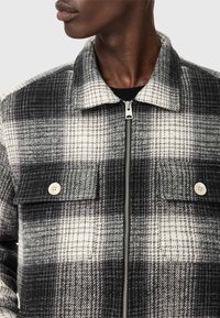 Veste à carreaux noir et blanc avec un col, fermeture éclair à l'avant et deux poches poitrine avec fermetures à bouton. Tissu doux et texturé.