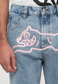 Jeans de mezclilla de color azul desgastado con un gran gráfico de un gato de dibujos animados rosa en el muslo izquierdo. Incluye bolsillos estándar y pasadores para cinturón.