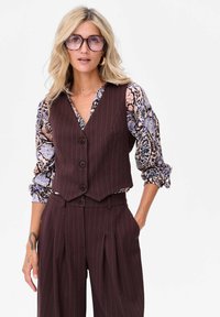 Brązowa kamizelka w paski, dopasowana do spodni z wysokim stanem, założona na blouse w kwiatowy wzór. Widać okulary i subtelny tatuaż.