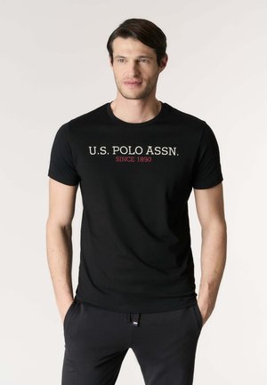 Uomo che indossa una maglietta nera U.S. Polo Assn. con scritte bianche e rosse, in piedi con le mani nelle tasche contro uno sfondo chiaro e uniforme.