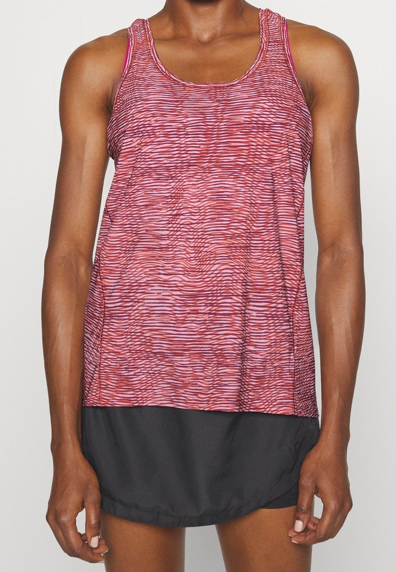 Femme portant un débardeur de sport sans manches rose et rouge à motifs, et une jupe noire, les mains relaxées sur les côtés.