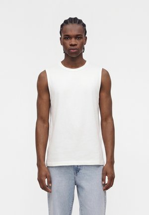 Young man with braided hair wearing a white sleeveless shirt and light blue jeans, standing against a plain light background.