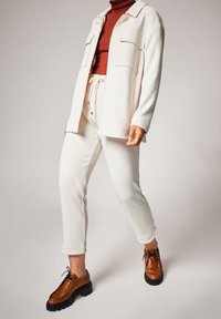 Veste texturée crème avec deux poches avant, associée à un pantalon de jogging crème assorti et des chaussures marron à lacets. La tenue a une coupe décontractée.