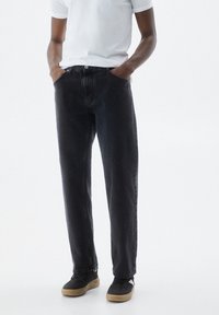 Jeans de mezclilla negra con un corte relajado, cinco bolsillos, costuras visibles y un diseño de pierna recta. Combinados con una camiseta blanca de manga corta.