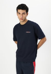 Sötétkék pamut póló rövid ujjakkal, kerek nyakkivágással, és egy kis hímes "GUESS JEANS" logóval a bal mellen. Simább textúra.