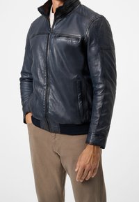 Man i en marinblå läderjacka med dragkedjefickor och beige byxor, stående med ena handen i fickan och den andra vid sidan.