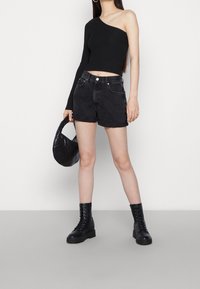 Schwarzes, geripptes Top mit einem Schulterträger, kombiniert mit hoch taillierten, schwarzen Denim-Shorts. Das Model trägt schwarze Schnürstiefel und hält eine schwarze, gesteppten Handtasche.