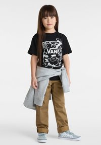 Schwarzes T-Shirt mit weißem grafischem Logo, khakifarbene Hose, grauer Pullover um die Hüfte gebunden und hellblaue Skate-Schuhe mit einem karierter Muster.