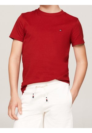 Barn klædt i en ensfarvet rød kortærmet t-shirt med lille logo og hvide shorts med snøre, stående med den ene hånd i lommen.