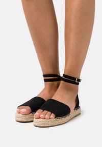 Černé semišové espadrilky se širokým páskem na prsty, páskem kolem kotníku se sponou a juty provázkovou podrážkou. Nohy jsou postavené vedle sebe.