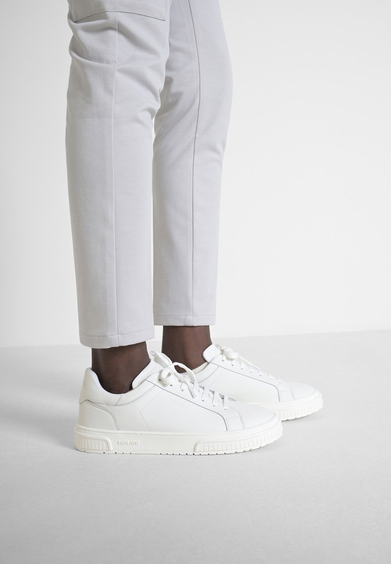 Sneaker bianche in pelle con punta rotonda, lacci, suola in gomma testurizzata e branding discreto. Abbinati a pantaloni aderenti grigio chiaro.