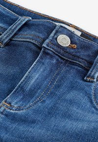 Primer plano de unos jeans de mezclilla azul mostrando el bolsillo delantero, los pasadores del cinturón y el botón de metal con la inscripción "Next Denim".