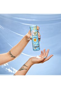 Tube de crème à motif floral bleu tenu au-dessus d'une paume ouverte, une noisette de crème visible. Texture lisse, finition brillante. Fond bleu clair.