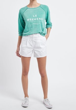 Personne portant un t-shirt vert à manches longues avec le texte « LE WEEKEND », un short blanc et des baskets blanches, debout devant un fond blanc uni.