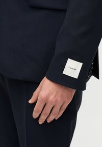 Λεπτομέρεια σακακιού σε ναυτικό μπλε, με λευκή τετράγωνη ετικέτα που γράφει "Calvin Klein", τοποθετημένη στο μανίκι κοντά στον μανσετί.