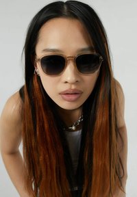 Bruna gradientsolglasögon med en genomskinlig båge. Modellen har rakt, långt svart och orange hår, och bär ett silverhalsband.