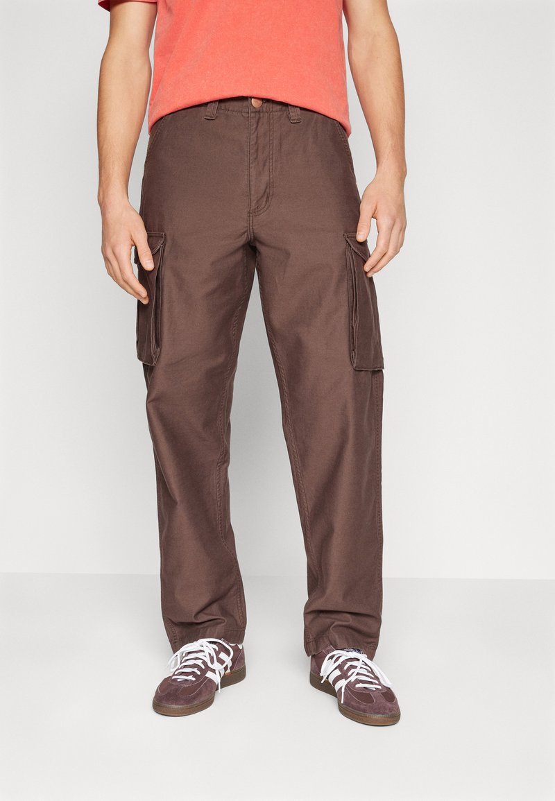Pantalones cargo marrones de algodón con bolsillos laterales, corte recto y una cintura con cierre de botón. Combinados con zapatillas oscuras.