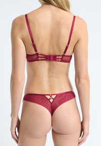 Femme portant de la lingerie transparente bourgogne avec des détails de lanières au dos du soutien-gorge et un string assorti avec des accents en métal, vue de derrière.