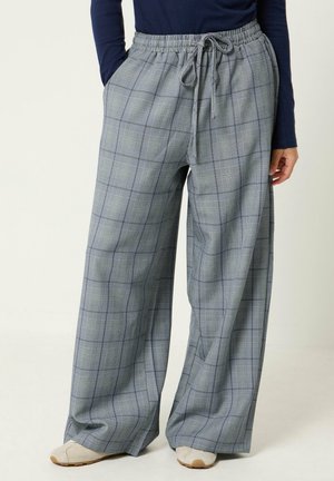 Lockere graue Karohose mit elastischem Bund und Kordelzug, getragen mit einem marineblauen Oberteil und beigen Schuhen, die Hände entspannt an den Seiten.